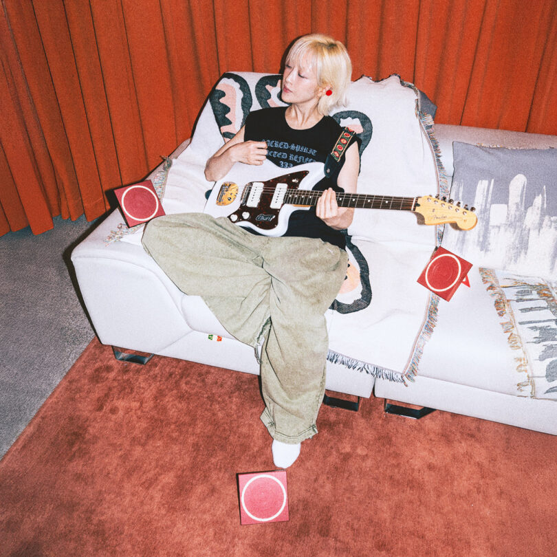 A person with short blonde hair sits on a white couch, playing an electric guitar, surrounded by four small red speakers, with orange curtains and a pink rug in the background.