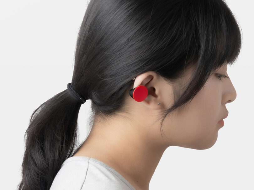 A person with dark hair in a ponytail wears a large, round, red earbud in their left ear against a plain white background.