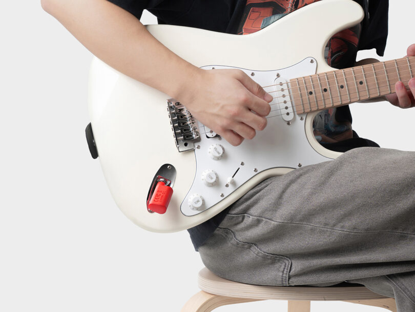 Person sitting on a stool playing a white electric guitar with a red lock attached to the bridge.