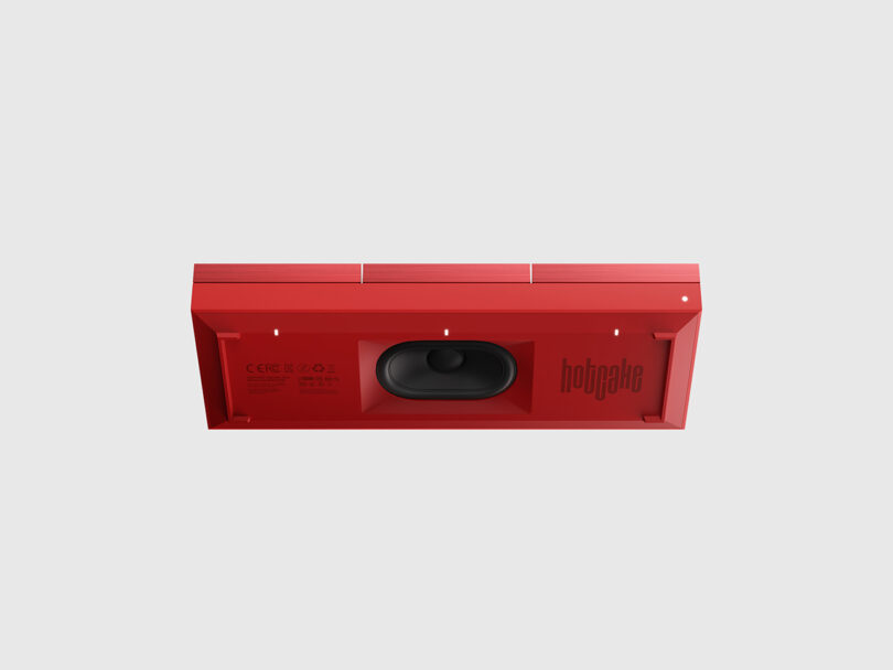 A rectangular red electronic device viewed from the bottom, showing a central speaker, ports, product information, and a logo on a light gray background.