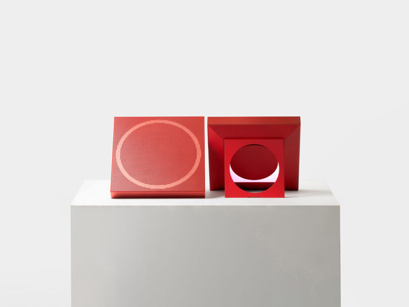 Two red geometric objects on a white pedestal; one is a square with a circular pattern, the other is a square frame with a circular cutout, both against a plain light background.