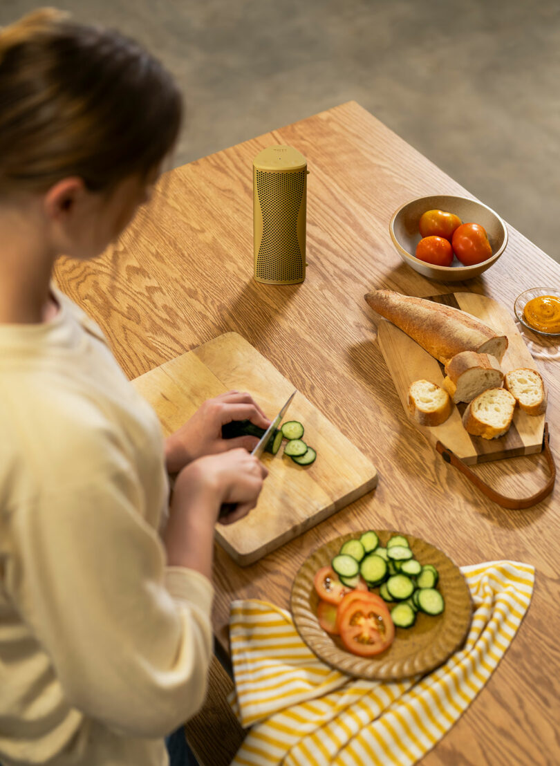 Person slicing cucumber on a wooden cutting board, with tomatoes, a baguette, and a yellow-striped cloth on a wooden table.