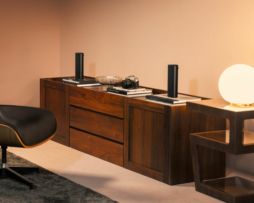 A wooden sideboard with drawers holds books, a camera, two black speakers, and a decorative bowl; a modern chair and a lit lamp on a side table are nearby.