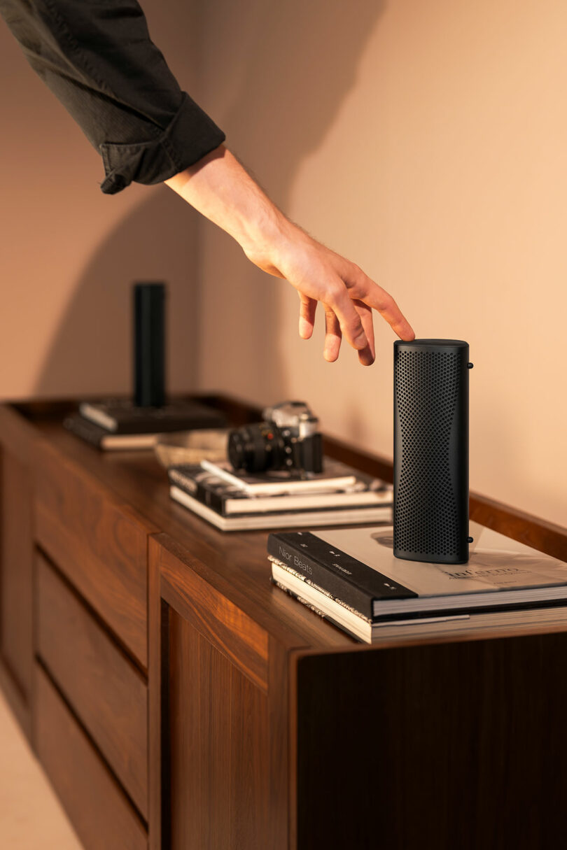 A person’s hand reaches to touch a black cylindrical speaker placed on a stack of books on a wooden sideboard, with a camera and another speaker in the background.