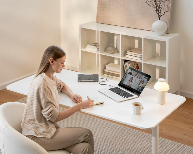 A woman sits at a white desk, writing in a notebook during a video call on her laptop. A cup, lamp, and speaker are also on the desk in a modern, minimalist room.