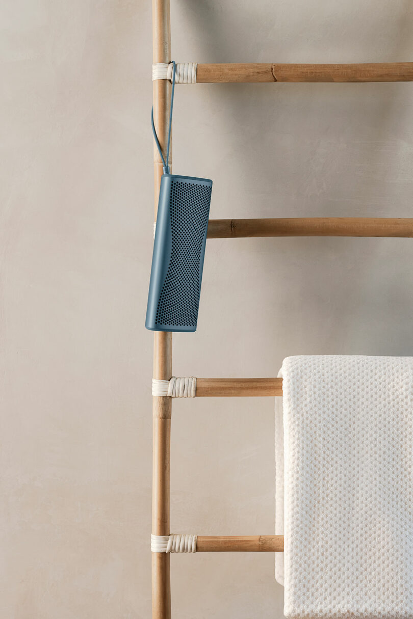 A blue portable speaker hangs from a rung of a wooden ladder, which also holds a folded white textured towel.