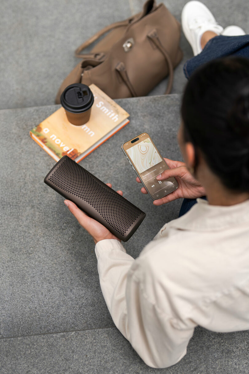 Person sitting on steps holding a smartphone and a portable speaker, with a brown bag, coffee cup, and a novel placed nearby.