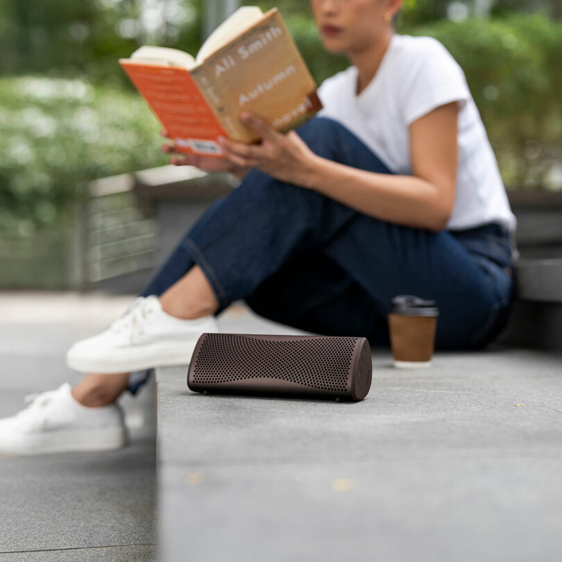A portable wireless speaker sits on a concrete step; in the background, a person in casual clothes reads a book and holds a cup of coffee.