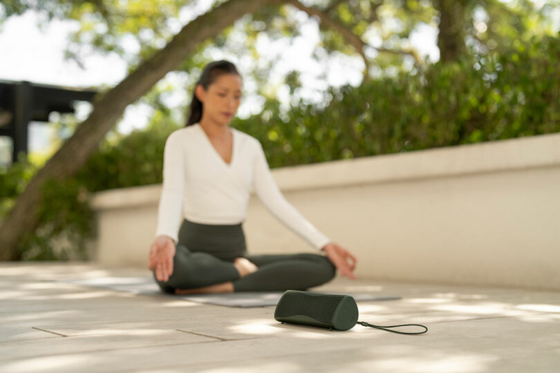 A woman sits cross-legged on a yoga mat outdoors, with a portable speaker placed on the ground in the foreground.
