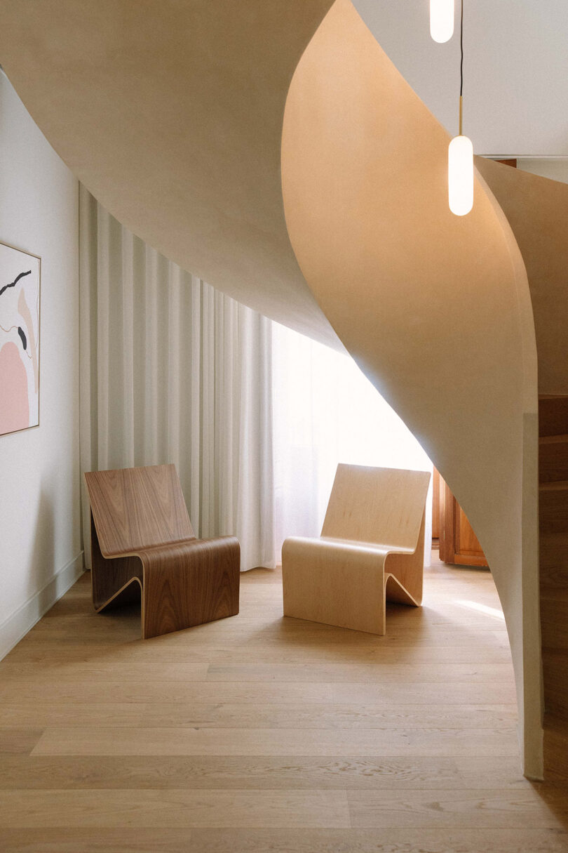 Modern interior with two minimalist wooden chairs beneath a curved staircase, light wood flooring, sheer curtains, pendant lights, and abstract wall art.