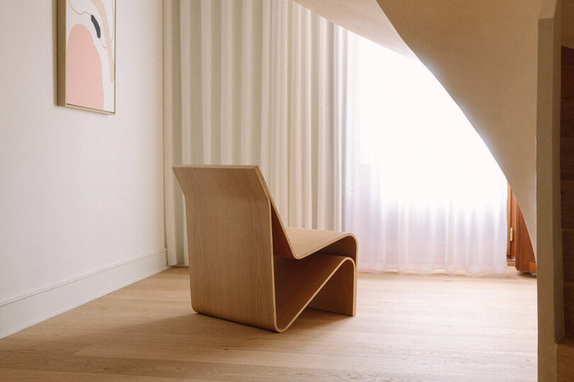 A minimalist room with a sculptural wooden chair, light wood flooring, sheer white curtains, and abstract wall art.
