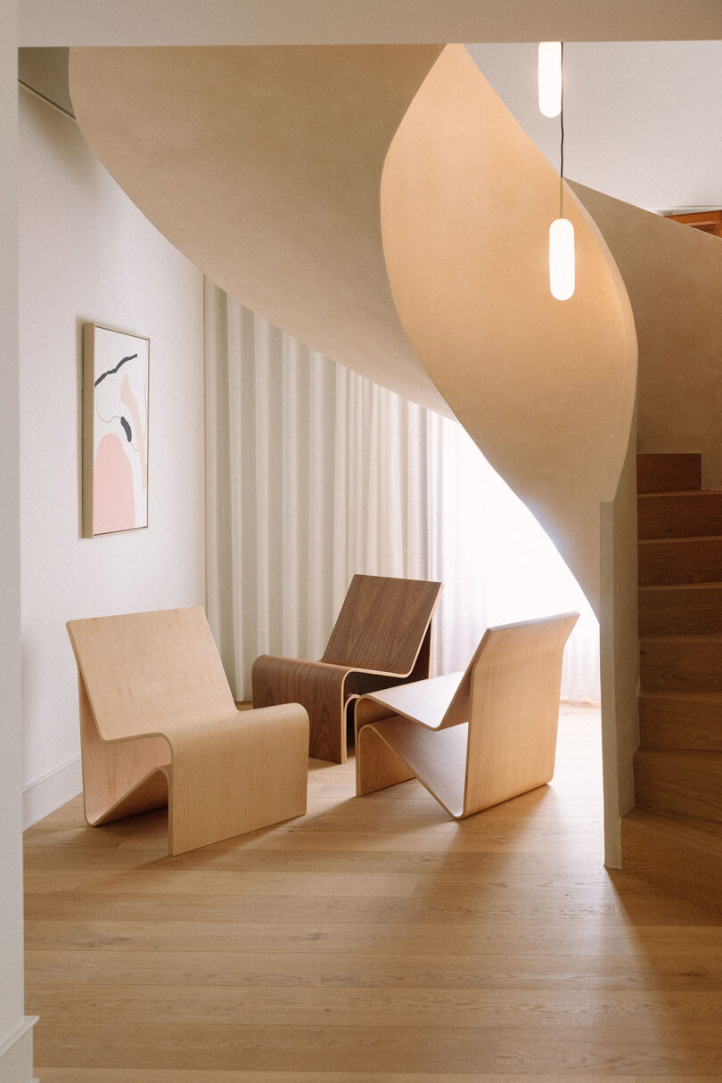 Three modern wooden lounge chairs are arranged under a curved staircase in a minimalist room with light wood floors, a hanging light, a curtain, and abstract wall art.