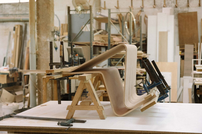 A bent plywood sheet clamped to wooden supports sits on a workbench in a cluttered woodworking shop.