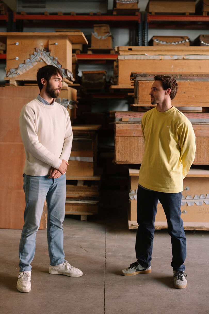 Two men stand facing each other in a warehouse with large wooden crates and boxes stacked behind them.