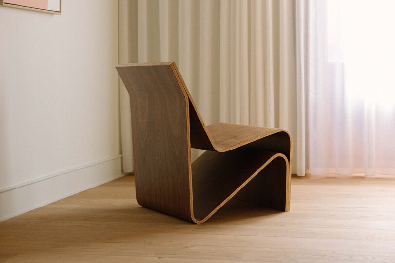 A sculptural wooden chair with a modern, angular design sits on a light wood floor near a window with sheer white curtains.