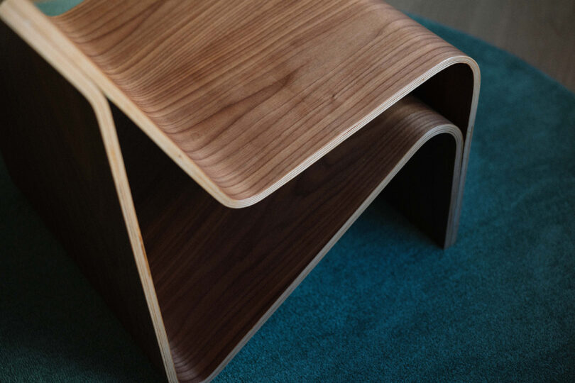 A wooden stool with a smooth, wavy design sits on a teal carpet.