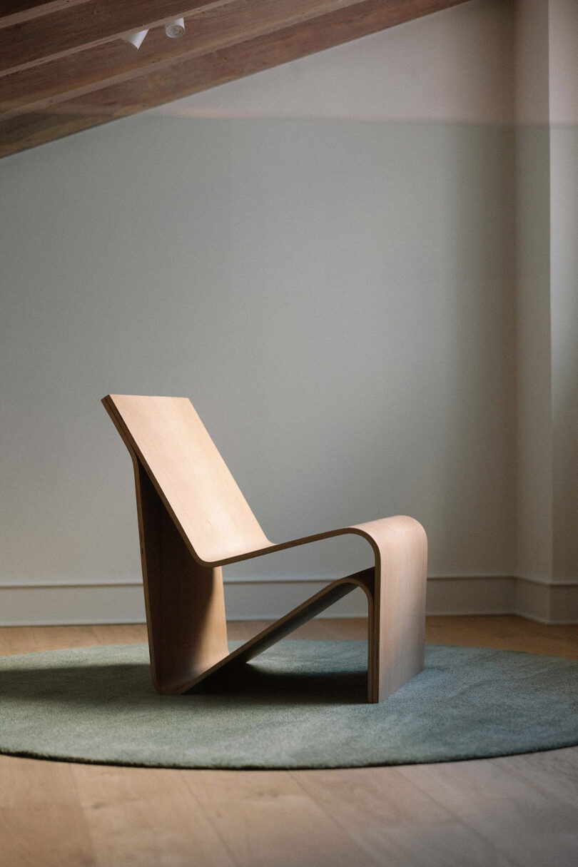 A modern wooden lounge chair with a minimalist, curved design sits on a round gray rug in a softly lit room.