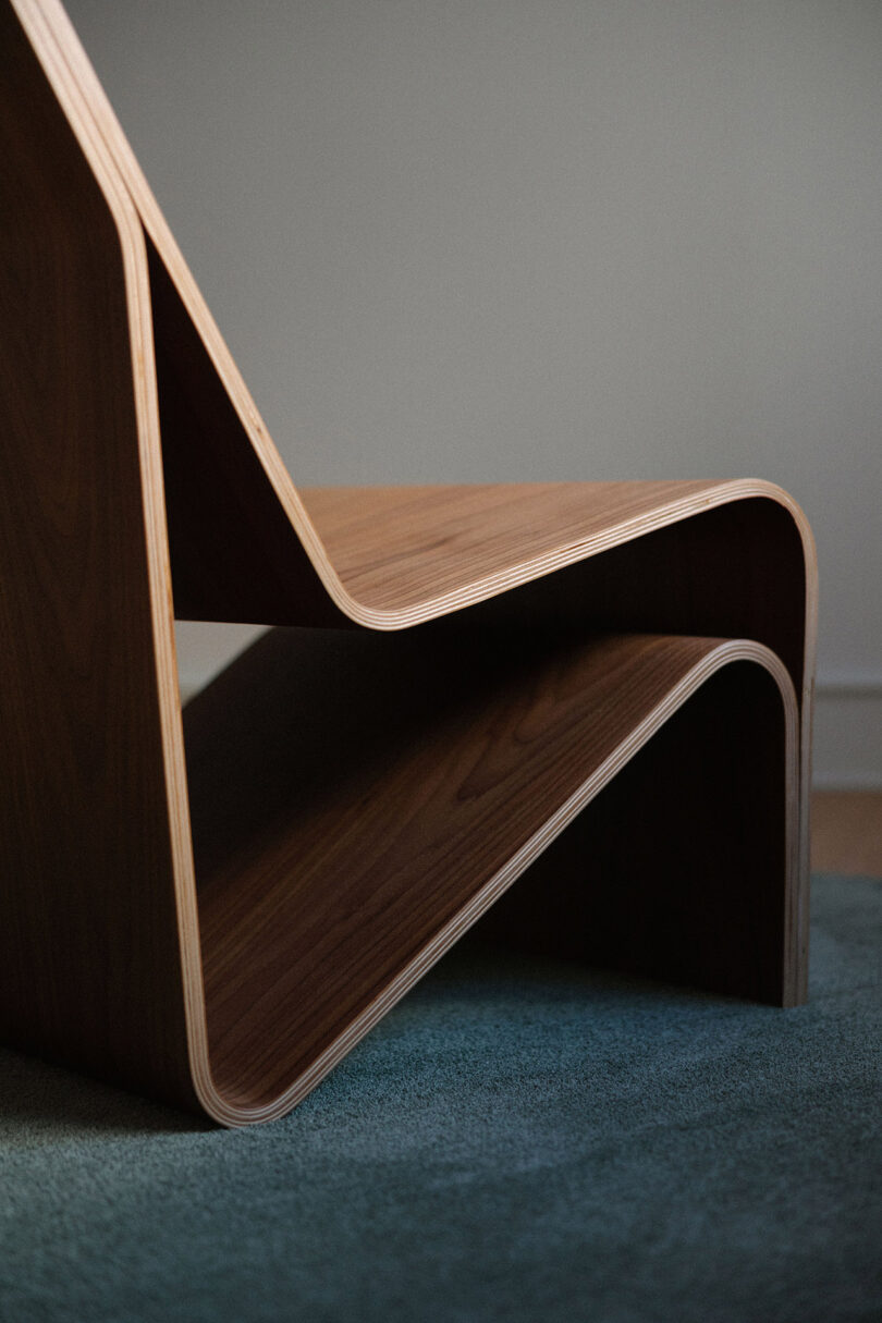 A close-up view of a modern wooden chair with curved, layered design placed on a textured blue carpet.