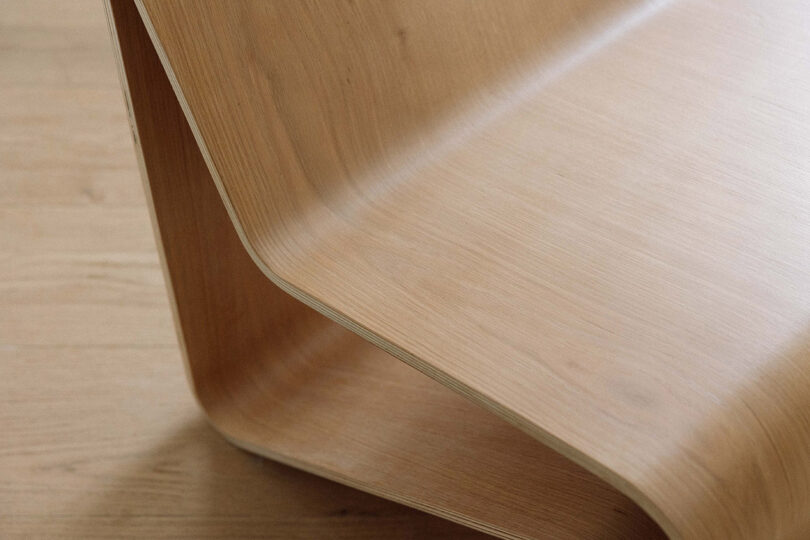 Close-up view of a modern, curved wooden furniture piece on a wooden floor, showing smooth, light-colored wood grain and angled edges.