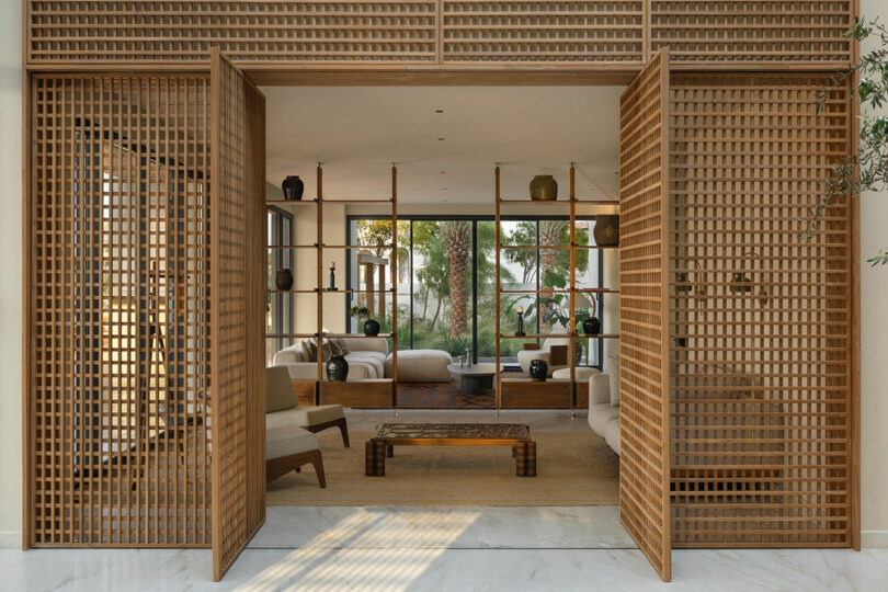 A modern living room with wooden lattice sliding doors, neutral-toned furniture, a low coffee table, and floor-to-ceiling shelving displaying vases and decor, with large windows showing greenery outside.