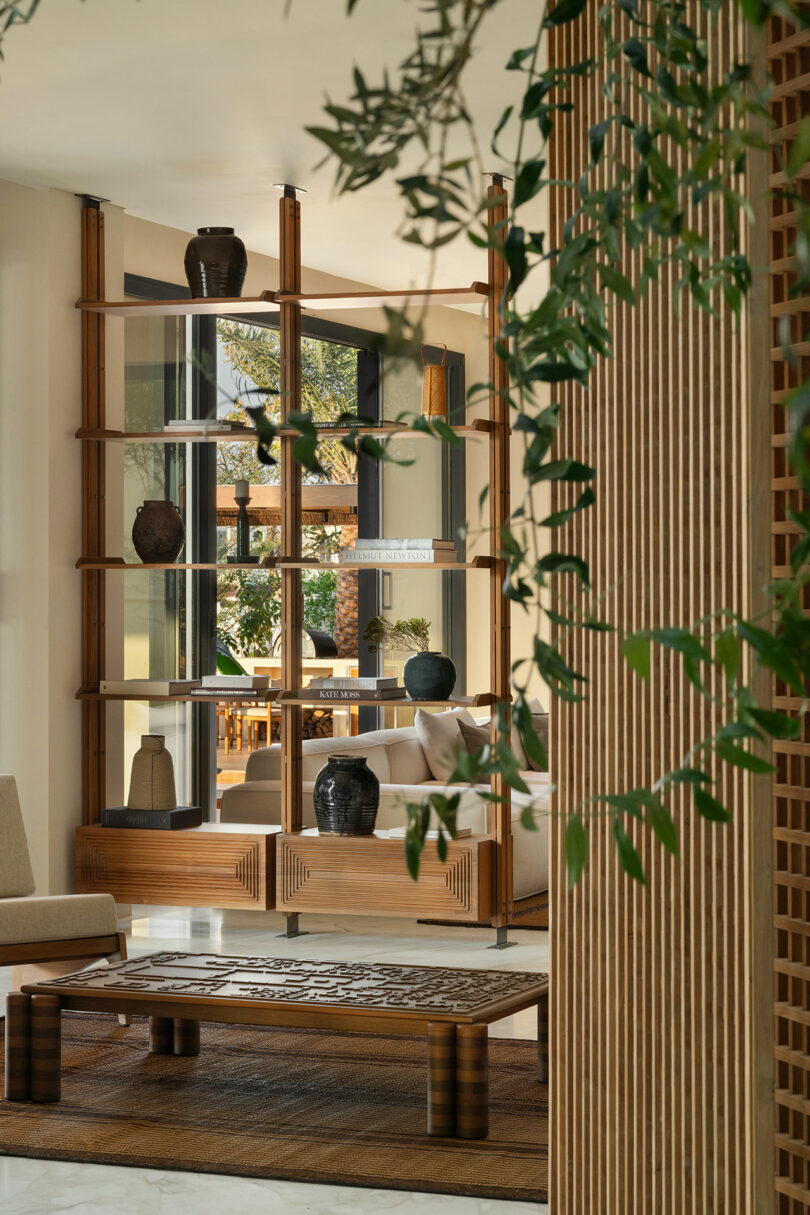 Modern living space with wooden shelving, ceramic vases, beige seating, and a wooden coffee table. Greenery partially visible in the foreground. Natural light fills the room.