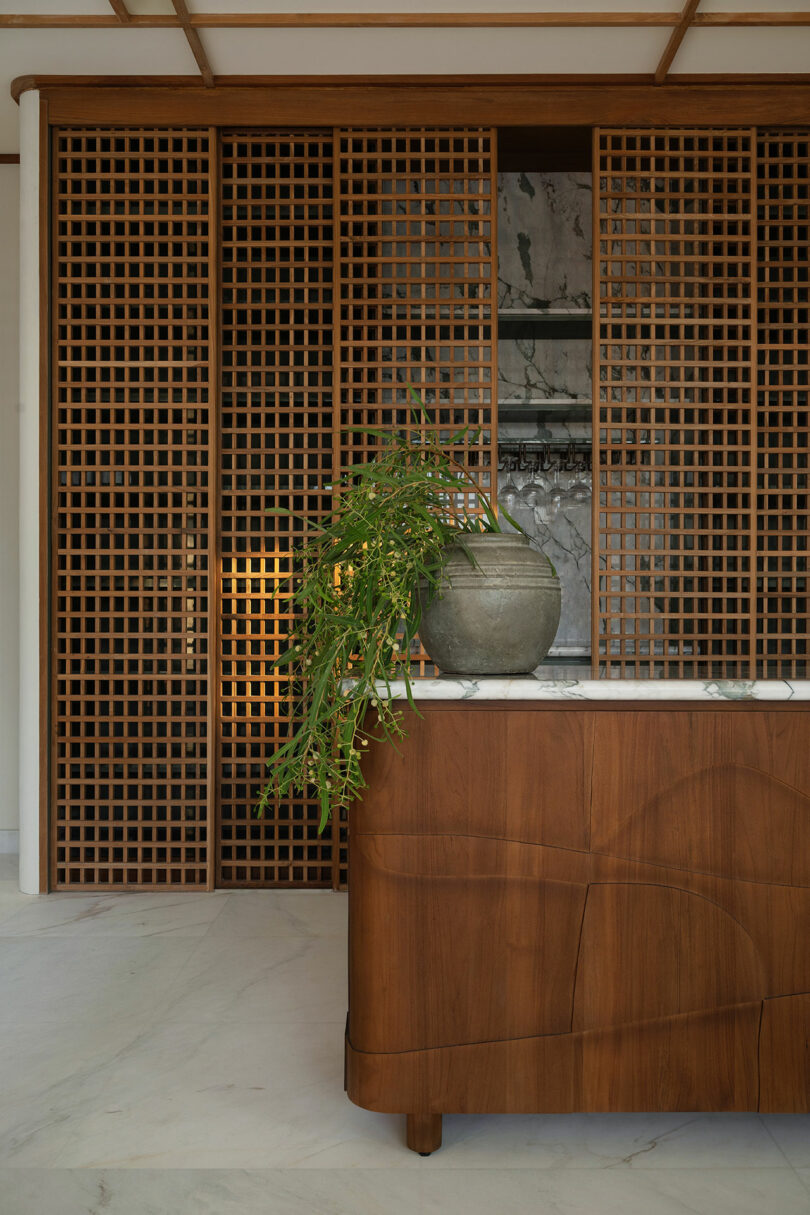 A wooden reception desk with a potted plant sits in front of a large lattice wood partition, with marble flooring and subtle interior lighting.