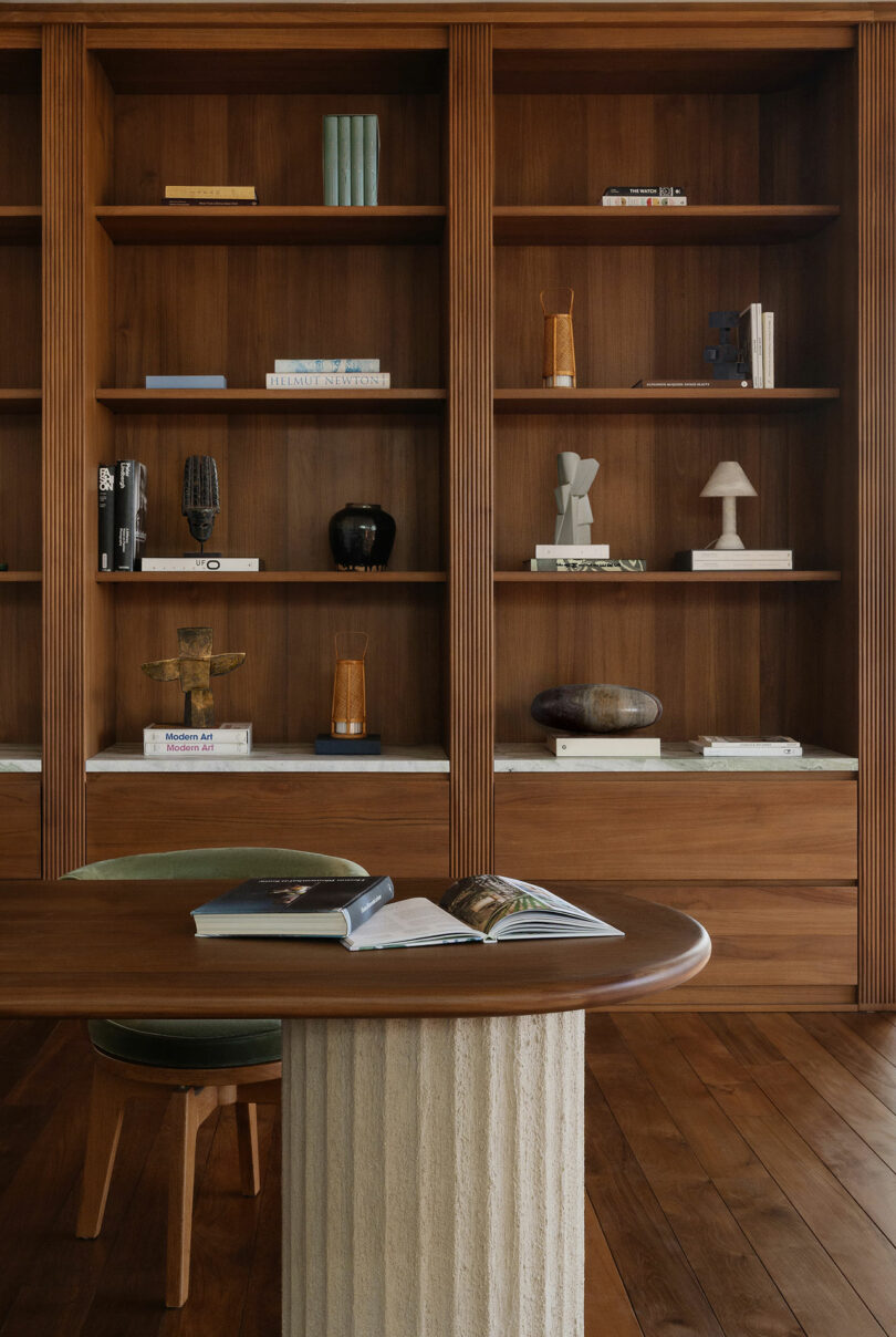 A round wooden table with an open book sits in front of built-in wooden shelves filled with books, decorative objects, and art pieces in a warmly lit room.