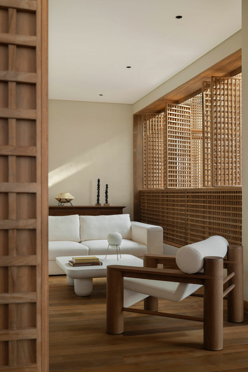 Minimalist living room with wooden furniture, white upholstered sofa and chair, coffee table with books, decorative items, and a wooden lattice screen along large windows.