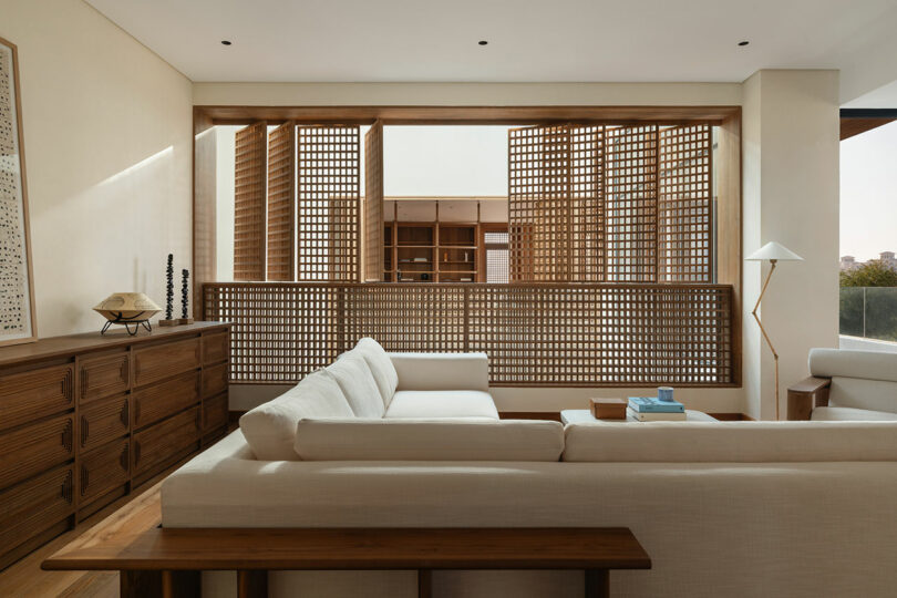A modern living room with a white sectional sofa, wooden furniture, and a wall of wooden lattice panels allowing natural light to enter.