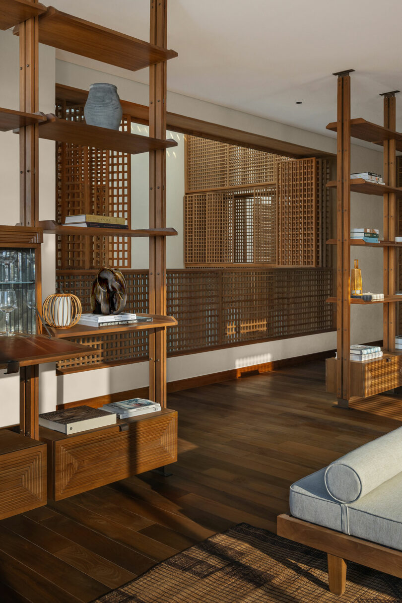 A modern living space with wooden shelving, decorative objects, books, a blue cushioned seat, and a wood slat partition.