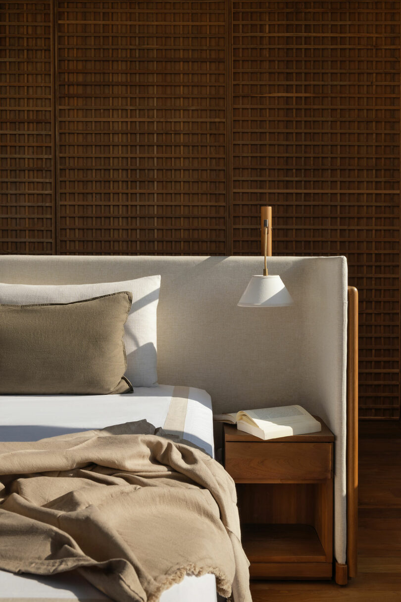 A neatly made bed with beige linens and a dark pillow beside a wooden nightstand holding an open book, with a wall-mounted lamp and a wooden accent wall in the background.