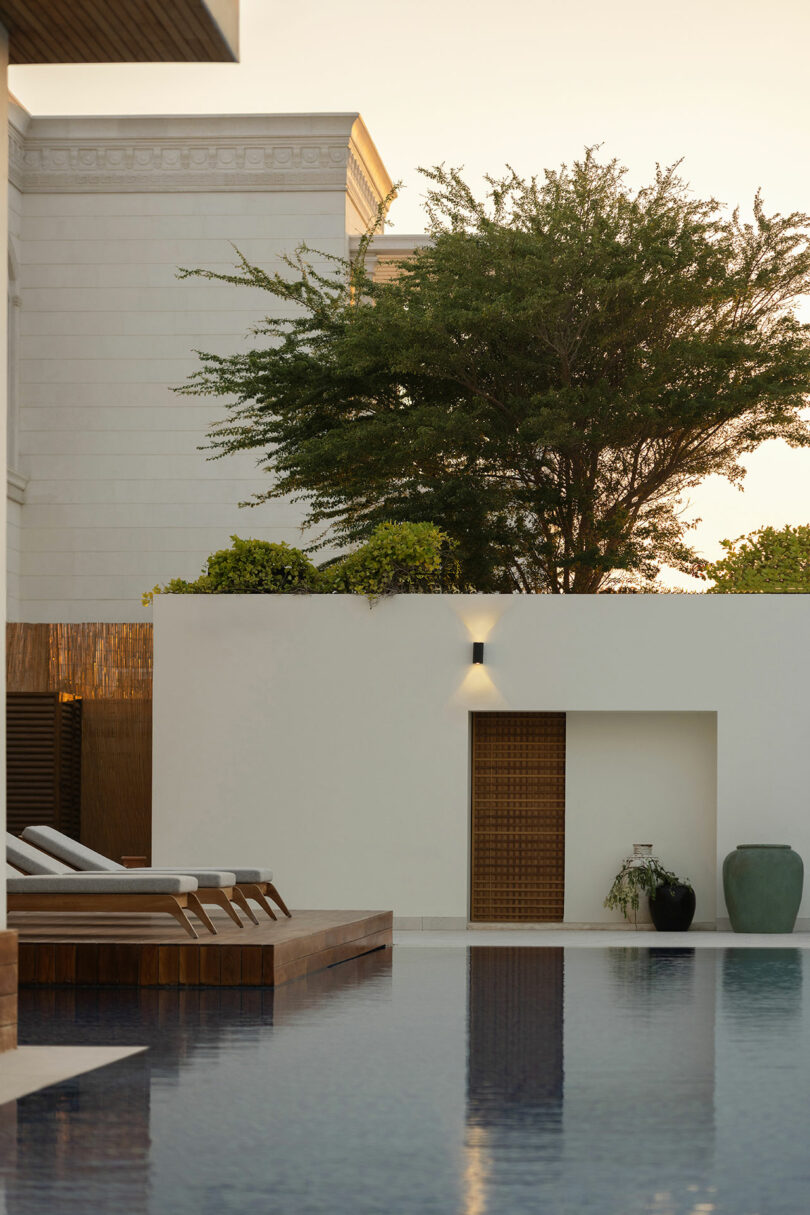 A serene outdoor pool with lounge chairs, a white wall, green trees, and decorative pots, photographed at sunset.