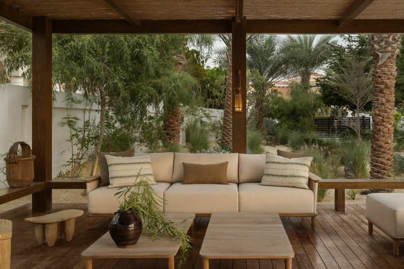 Outdoor patio with a beige cushioned sofa, striped pillows, wooden tables, and lush green plants and trees in the background.