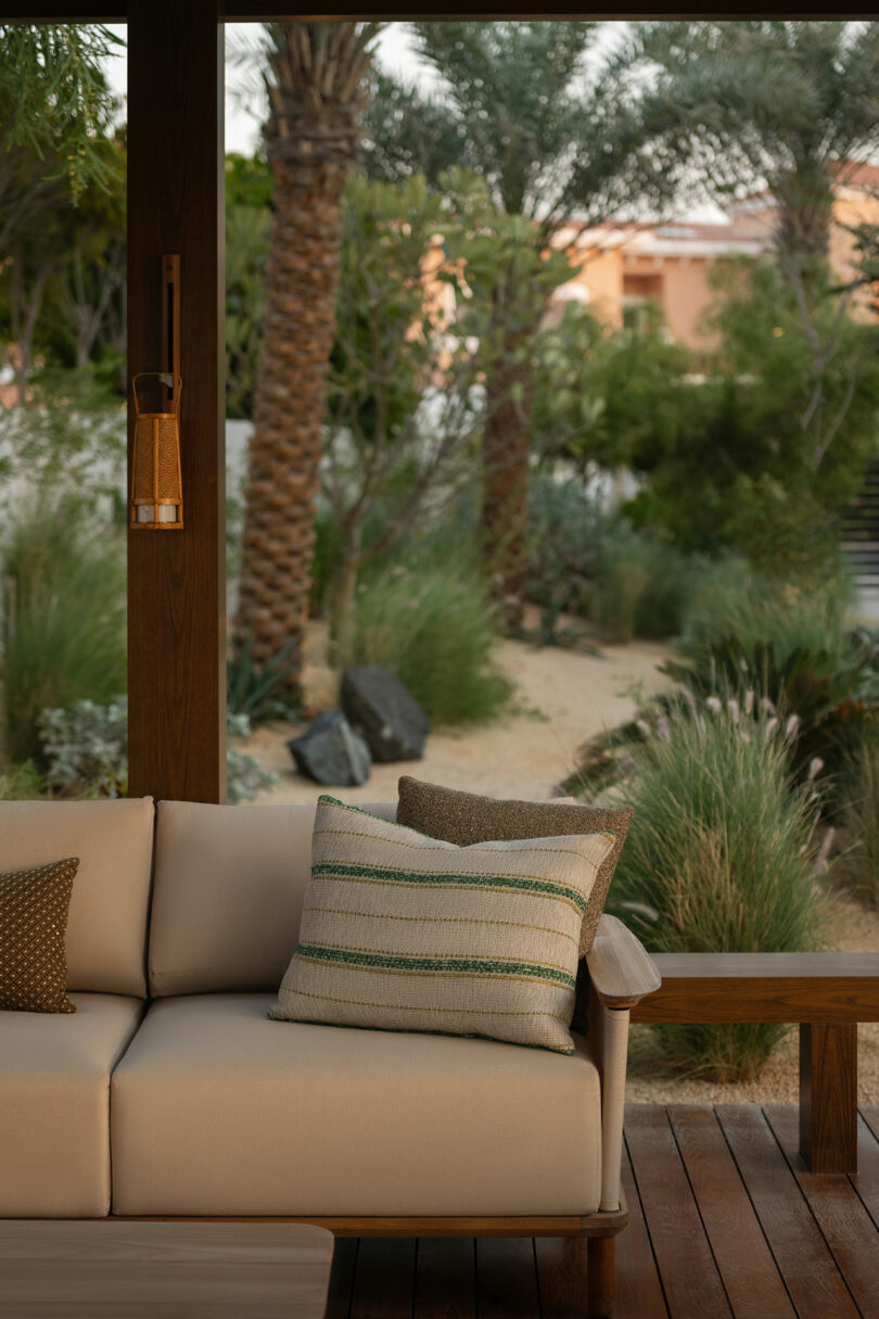 Outdoor patio with a beige sofa and striped pillow, wooden floor, bench, and lush garden with palm trees and greenery in the background.