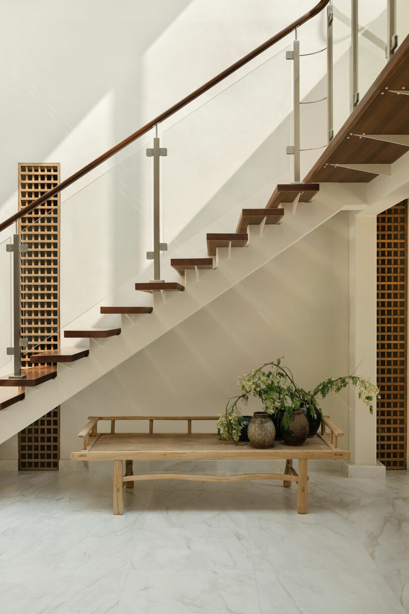 A modern wooden staircase with glass railing above a minimalist bench holding potted plants, set against a white wall and marble floor.