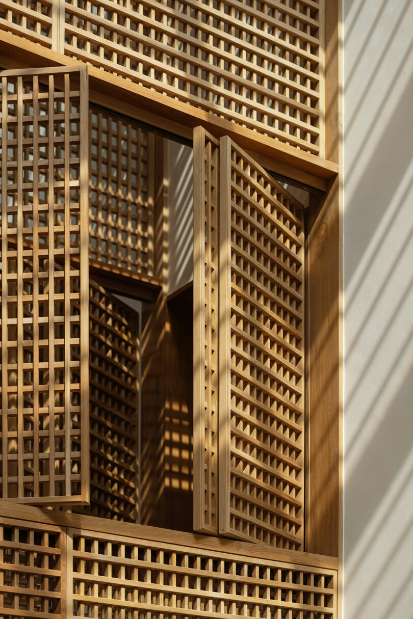 Wooden lattice panels arranged in a geometric pattern, with some panels partially open, casting grid-like shadows on a light-colored wall.