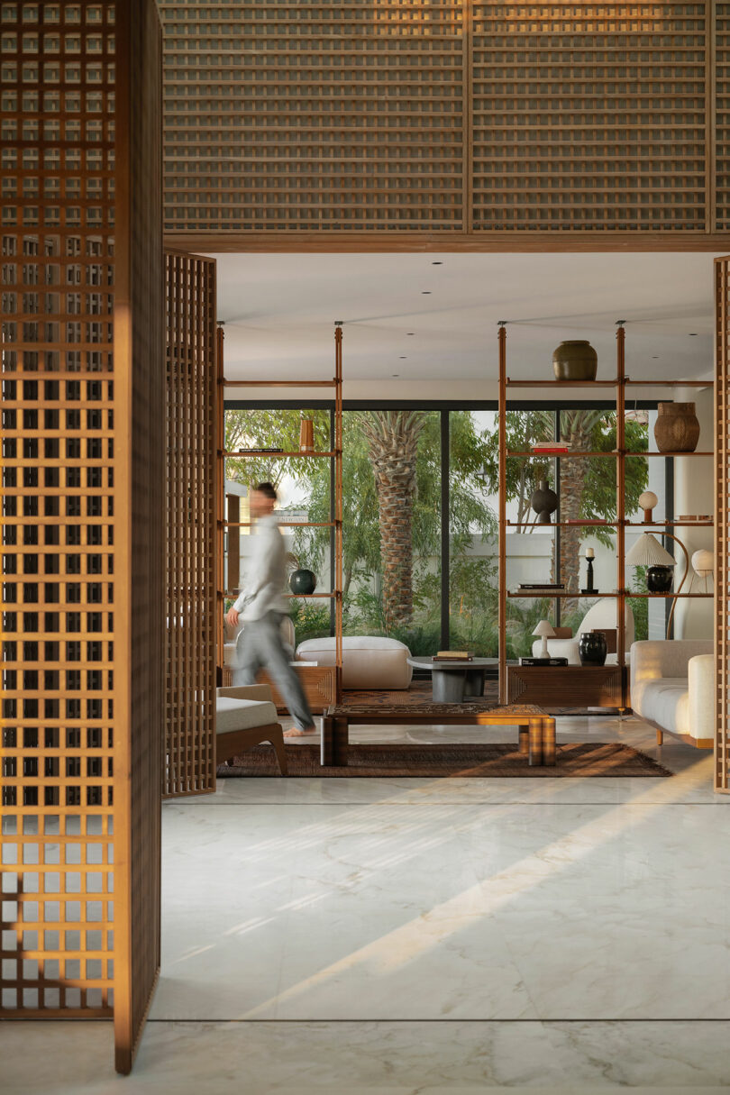 A person walks through a modern, sunlit lounge with wooden lattice partitions, open shelving, neutral-toned furniture, and large windows overlooking palm trees.