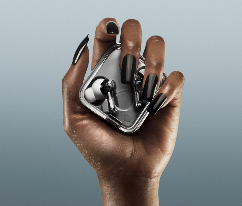 Una mano con uñas negras largas sostiene una pequeña caja de metal que contiene un par de tapones para los oídos inalámbricos y sobre un fondo normal.