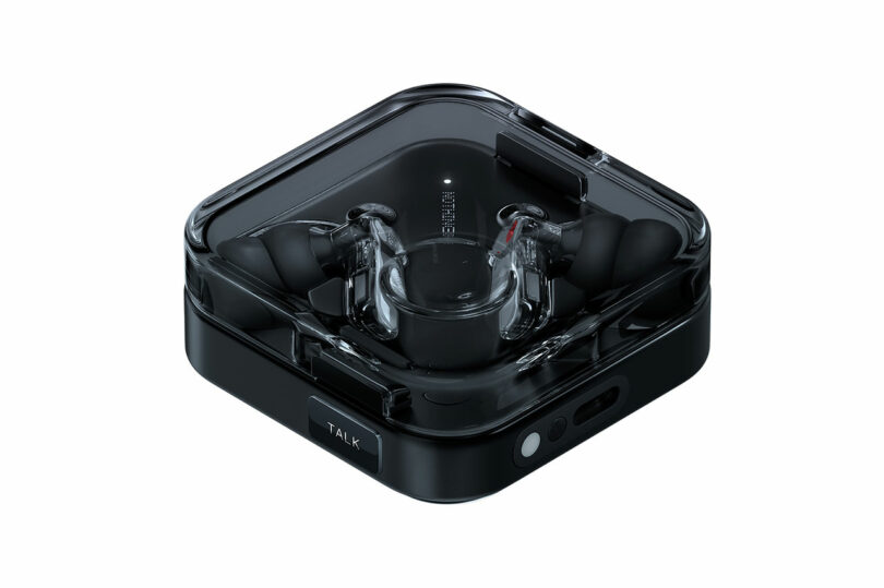 Un par de auriculares inalámbricos negros se colocan en una caja de carga cuadrada transparente con electrónica visible y botones de esquina marcados.