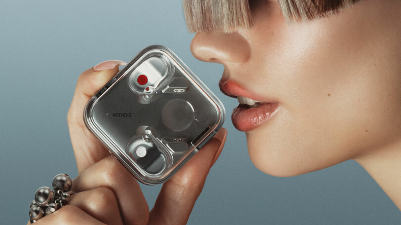 Un hombre con golpes cortos contundentes contiene un dispositivo electrónico cuadrado de metal con botones y un punto rojo en la boca cerca de un fondo plano.