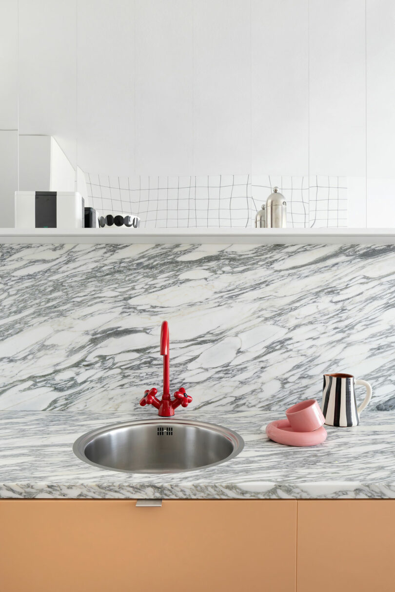 Modern kitchen with a marble countertop and backsplash, a round stainless steel sink, a red faucet, pink dishes, a striped jug, and minimalist decor on a white shelf.