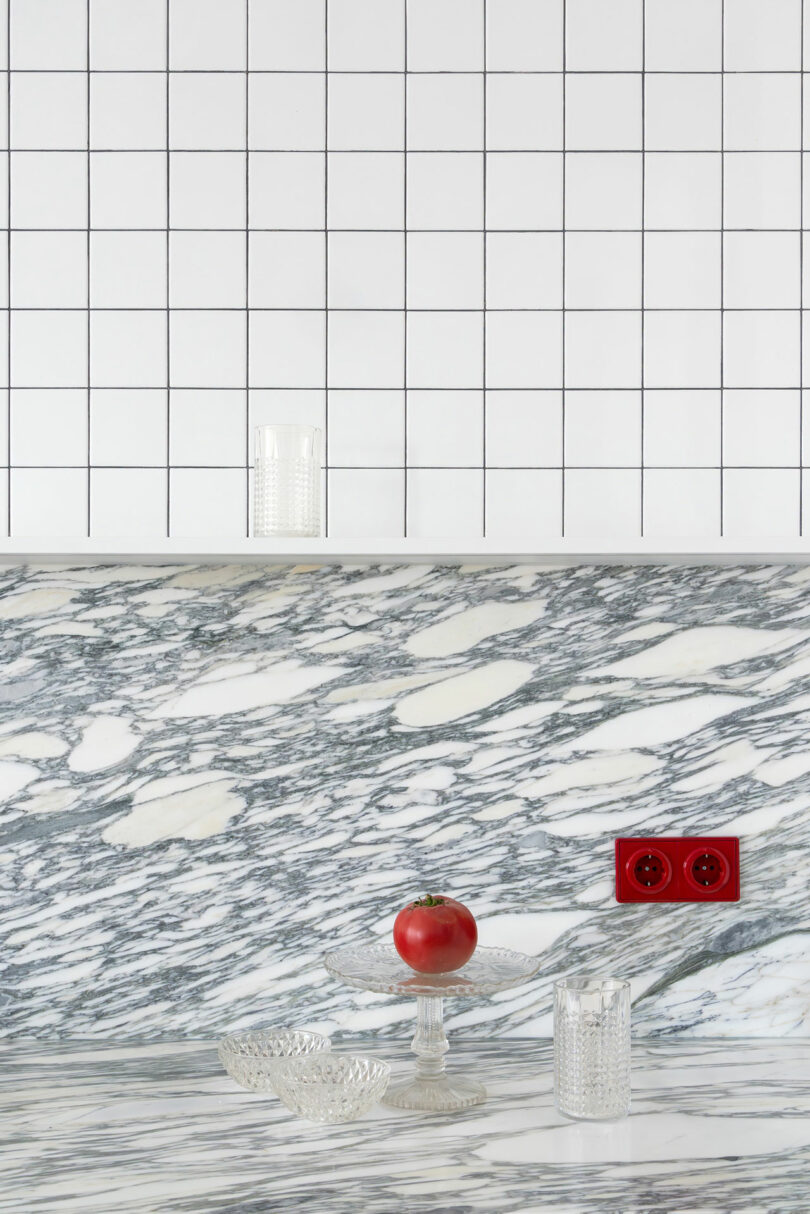 A tomato and two clear glass cups are placed on a marble countertop with a red electrical outlet; white tiled wall is in the background.
