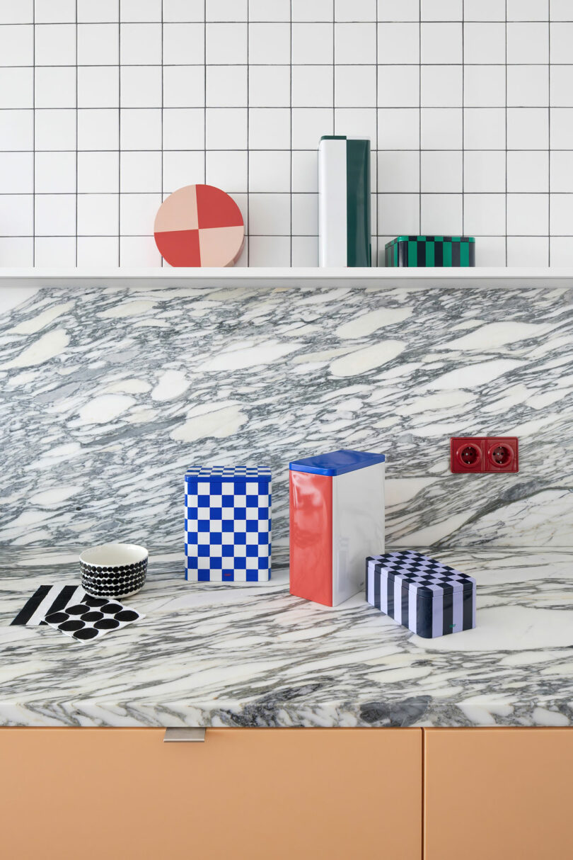 A marble countertop with geometric containers and bowls, a white grid tile backsplash, and a shelf holding books and a round object with a red and beige pattern.
