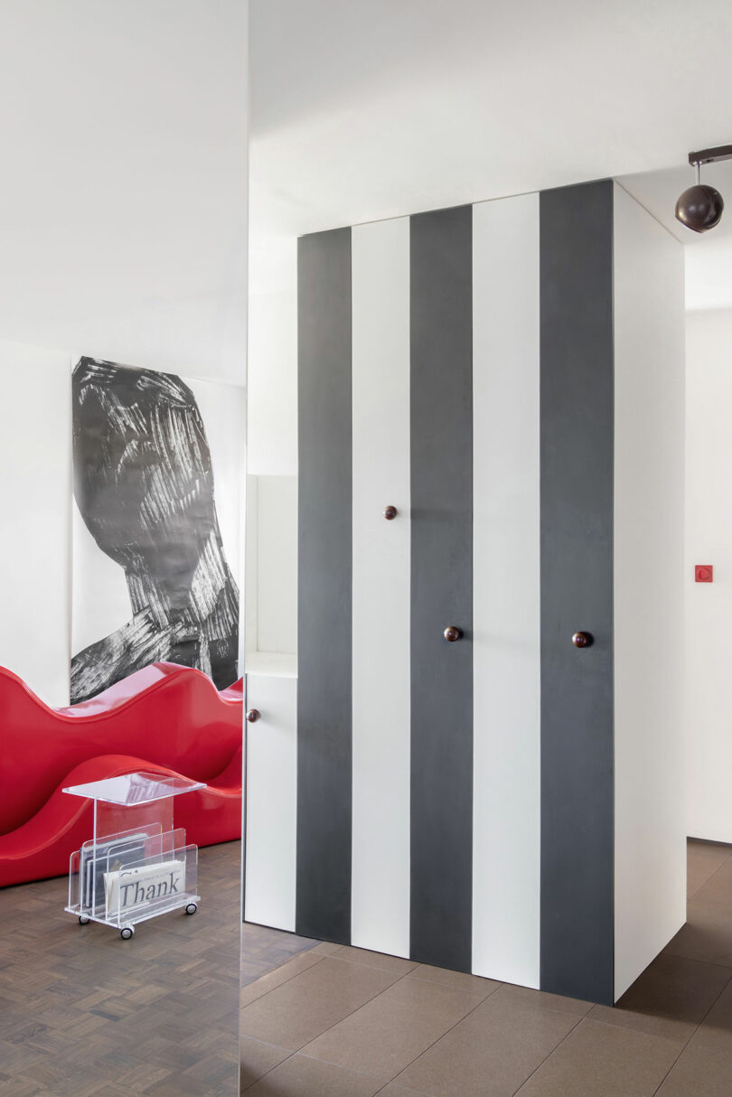 A modern interior with a red sculptural sofa, a striped black-and-white cabinet, abstract wall art, and a clear acrylic magazine holder on a tile floor.