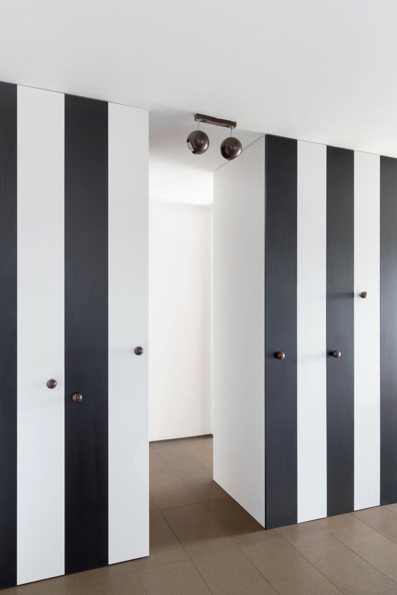 A hallway with floor-to-ceiling black and white vertical striped panels, brown door handles, and a ceiling light fixture with two round shades.