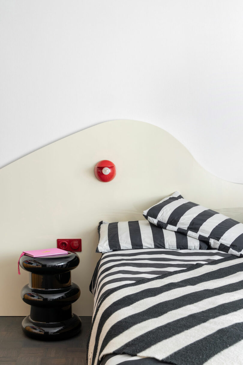 A bed with black and white striped bedding is next to a glossy black side table and a red wall sconce, set against a white wavy headboard and wall.
