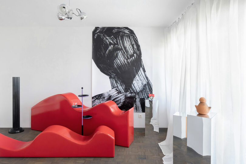 A modern room with wavy red seating, a black-and-white abstract artwork on the wall, white curtains, and minimalist sculptures on pedestals.
