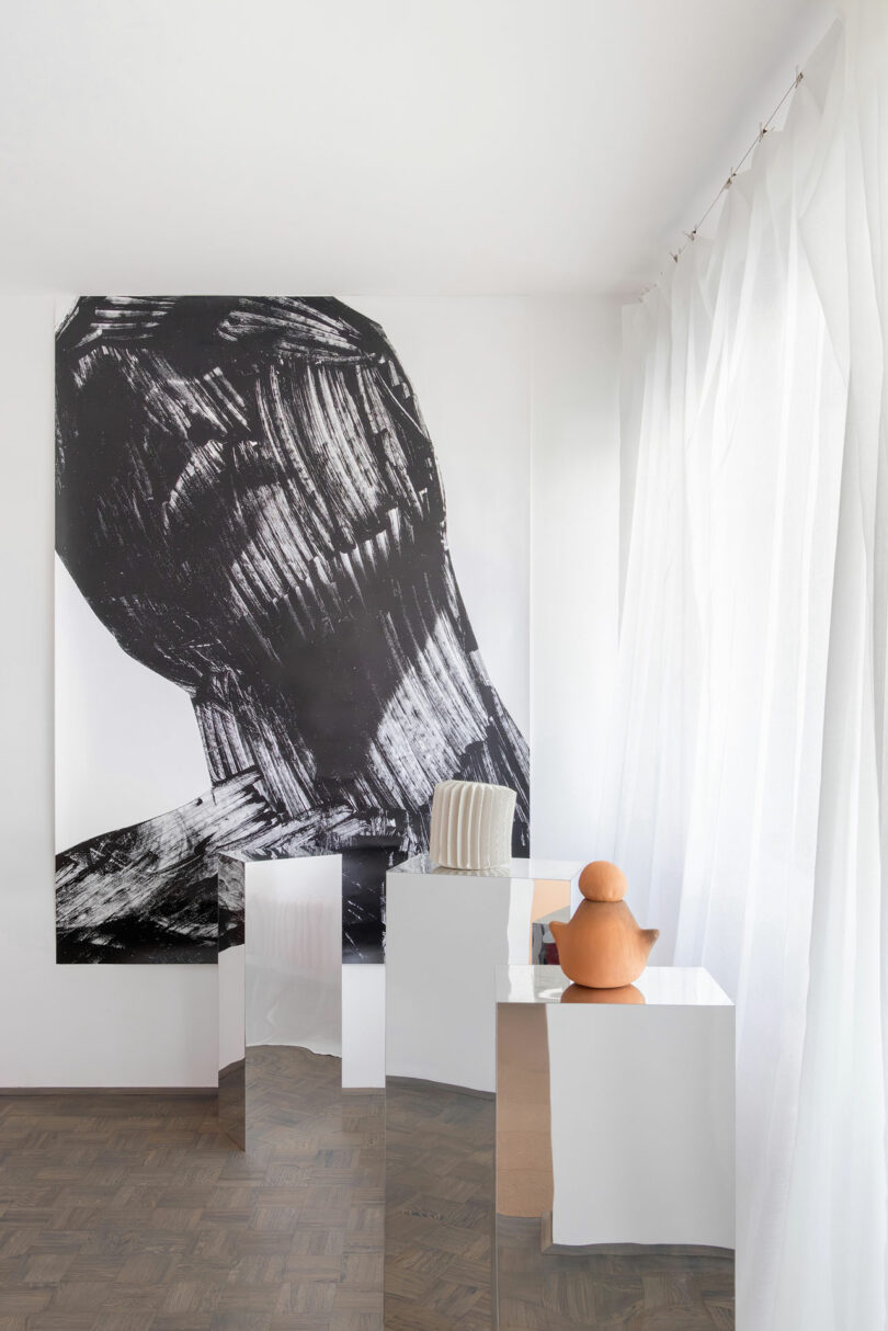 A minimalist room features a large abstract black-and-white artwork, mirrored pedestals, a striped pillow, a terracotta duck sculpture, and sheer white curtains.