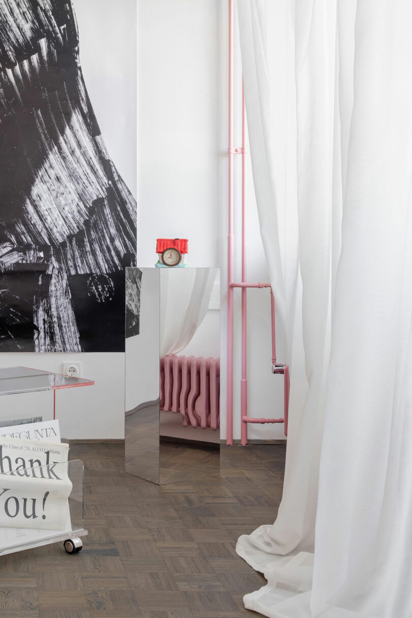 A mirrored pedestal with a red vintage clock stands next to pink radiator pipes, a curtain, and a cart with a transparent bag labeled “thank you!” in a modern room.