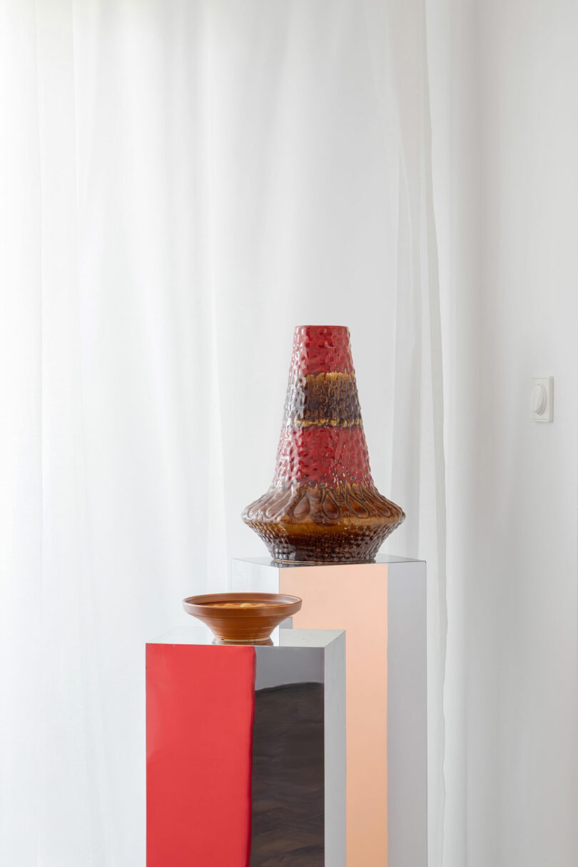 A textured, brown and red vase and a shallow brown bowl are displayed on two white pedestals against a white background with sheer curtains.